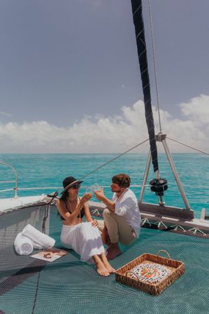 Couple toasting with wine on a catamaran trampoline above clear turquoise tropical waters, sunlit sky, picnic basket and rolled towels for a relaxed sailing getaway.