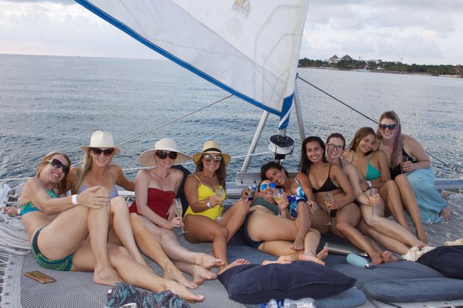 Group of friends lounging on the netting of a catamaran sailboat at sea, wearing swimsuits, sun hats and sunglasses, holding cocktails and smiling with a coastal shoreline in the background.
