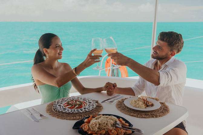 Romantic couple clinking wine glasses during a yacht lunch with turquoise tropical ocean view, plates of seafood, salad and mashed potatoes
