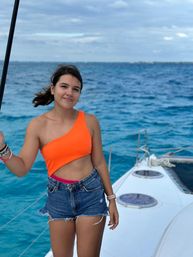 Smiling woman in an orange one-shoulder top and denim shorts standing on a sailboat deck with turquoise tropical ocean and cloudy sky — sailing vacation vibe