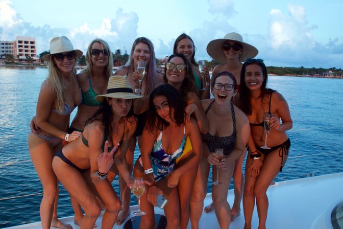 Group of ten women in bikinis on a boat near a tropical coastline at sunset, laughing and posing with champagne glasses and sun hats.