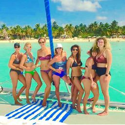 Seven women in colorful swimsuits posing on a catamaran deck with a blue-striped towel, clear turquoise water, palm-fringed sandy tropical beach and sunny sky in the background