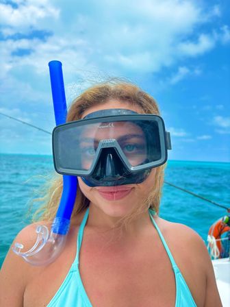 Close-up of person wearing a black snorkel mask and bright blue snorkel on a boat over turquoise tropical ocean under a bright blue sky, ready to snorkel.