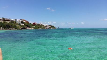 Tropical coastline with clear turquoise water, rocky shore lined with colorful houses, a distant pier, and an orange life buoy tethered floating in the foreground under a sunny blue sky.