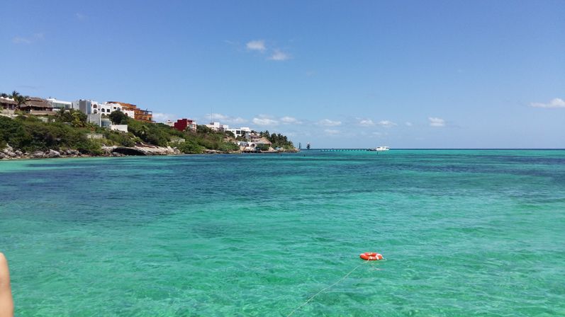 Tropical coastline with clear turquoise water, rocky shore lined with colorful houses, a distant pier, and an orange life buoy tethered floating in the foreground under a sunny blue sky.