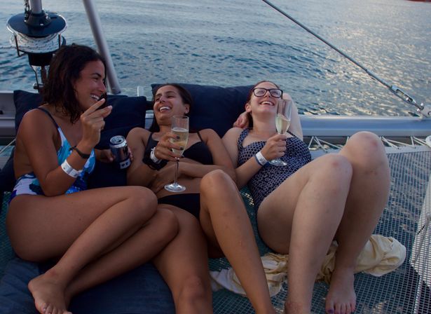 Three people in swimsuits laughing and lounging on a catamaran's net over calm ocean water, sipping champagne and a canned drink — relaxed sailing getaway.
