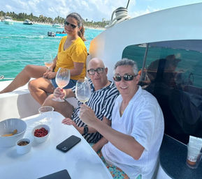 Three adults smiling and toasting with wine glasses on a white catamaran cruise near a palm-lined tropical coastline and turquoise water, with snack bowls on the table.