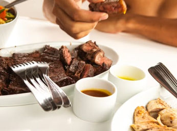 Close-up of a juicy grilled steak platter — hands picking up a steak bite from sliced steak on a white serving dish, with two dipping sauces, forks, grilled chicken slices and a bowl of mixed vegetables on a bright dining table.