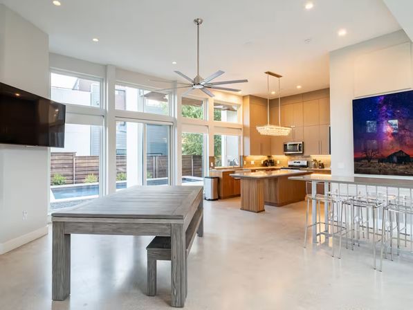 Bright modern open-concept kitchen and dining area with floor-to-ceiling windows overlooking a backyard pool, wood island with pendant chandelier, stainless appliances, ceiling fan, gray farmhouse table and clear acrylic bar stools