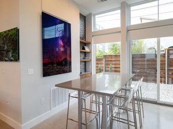 Bright contemporary dining nook with glossy island table and clear acrylic bar stools, tall windows and sliding glass door to a wooden-fenced urban patio, colorful starry-sky wall art and built-in shelving.