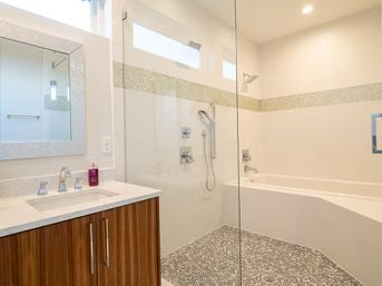 Bright modern bathroom with frameless glass walk-in shower and mosaic pebble floor, white soaking tub, wood-grain vanity with quartz countertop, and horizontal glass-tile accent strip.
