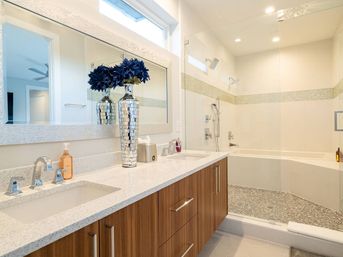 Bright modern master bathroom with double quartz vanity and wood cabinets, decorative silver vase, and glass-enclosed walk-in shower with mosaic tile — spa-like design.