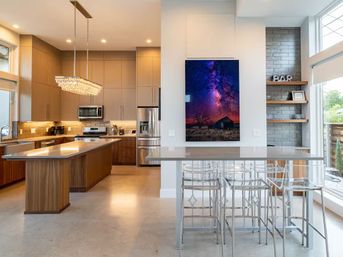 Bright modern open-concept kitchen with wood island and quartz countertops, crystal pendant chandelier, stainless steel appliances, breakfast bar with clear acrylic stools, large windows and framed Milky Way artwork.