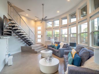 Sunlit contemporary living room with curved bank of tall windows, floating black staircase, gray sofas with teal and yellow pillows, round white coffee tables and oversized ceiling fan.