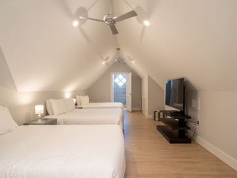 Bright attic loft bedroom with three beds in a row, crisp white linens, sloped ceilings with ceiling fans, bedside lamps, wall-mounted TV and light hardwood floors