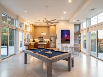 Bright, sleek open-concept game room and kitchen featuring a navy-felt pool table, bar seating, stainless-steel appliances, pendant lights, ceiling fan, and floor-to-ceiling windows.