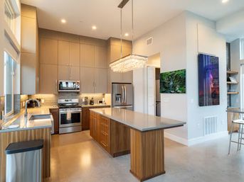 Bright modern open-concept kitchen in a contemporary home with a long quartz-topped island, wood lower cabinets and tall neutral upper cabinets, stainless steel range, fridge and microwave, crystal pendant chandelier, tile backsplash and polished concrete floor.