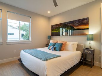 Bright modern master bedroom with white bedding, teal throw and pillows, fluffy pink accent cushions, panoramic mountain-sunset wall art, matching teal lamps on dark nightstands and a large window with roller shade