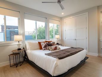 Sunlit modern city apartment bedroom with large windows, white bedding, rust-accent pillows, wooden nesting nightstands with lamps, ceiling fan, and light hardwood floors.