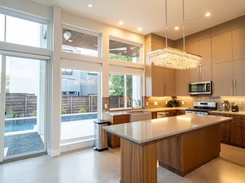 Sunlit contemporary kitchen with large windows and sliding glass door overlooking a backyard pool, wooden island with light quartz countertop, crystal pendant chandelier, stainless steel appliances, farmhouse sink, and warm recessed lighting