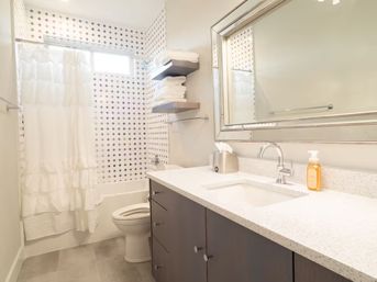 Bright, airy modern bathroom with a white shower-tub and black-and-white patterned tile accent wall, gray vanity with speckled quartz countertop, large framed mirror, floating shelves stacked with white towels, and a soap dispenser.