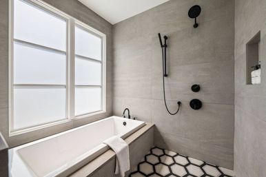 Modern minimalist bathroom with a white soaking tub under frosted windows, matte gray tile walls, black rain and handheld shower fixtures, and black-and-white hexagon floor tiles.