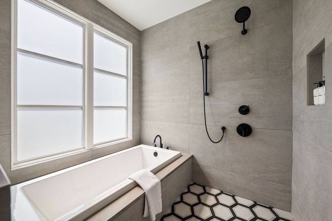 Modern minimalist bathroom with a white soaking tub under frosted windows, matte gray tile walls, black rain and handheld shower fixtures, and black-and-white hexagon floor tiles.