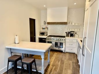 Bright modern white kitchen with marble-top farmhouse island, stainless-steel gas range, subway-tile backsplash, open shelves and hardwood floors