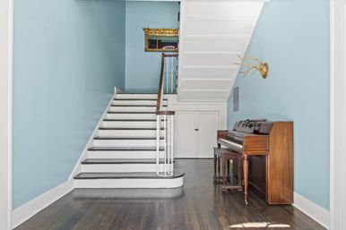 Light-blue stair hall with white wooden staircase, dark hardwood floors, under-stair storage doors, upright wooden piano with bench, gold antler wall mount and small gilded mirror.