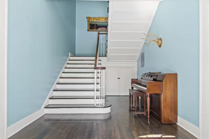 Light-blue stair hall with white wooden staircase, dark hardwood floors, under-stair storage doors, upright wooden piano with bench, gold antler wall mount and small gilded mirror.