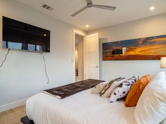Cozy modern bedroom with white bedding and colorful accent pillows, wall-mounted TV with visible cords, ceiling fan, open door and panoramic sunset landscape artwork