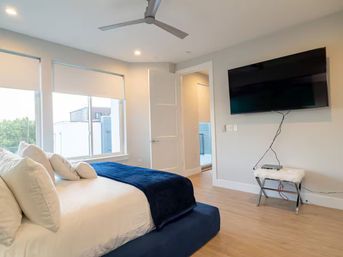 Modern bright master bedroom with white linens and navy throw on a low platform bed, large windows with roller shades, ceiling fan, wall-mounted flat-screen TV, light wood floors, and an open doorway.