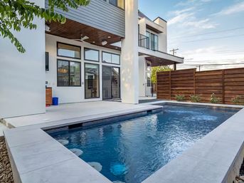 Modern backyard with rectangular plunge pool and concrete deck, covered patio with ceiling fan and large glass doors of a two-story contemporary home and wooden privacy fence