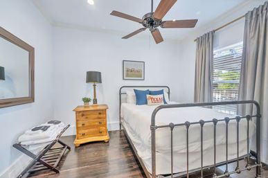 Bright modern bedroom with black metal bed frame and white linens, TEXAS accent pillow, wooden nightstand with lamp, ceiling fan, dark hardwood floor, and window with blinds and gray curtains.