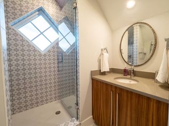 Bright modern bathroom with glass walk-in shower lined in geometric patterned tile and a diamond-shaped window, wood vanity with beige countertop, round framed mirror and chrome fixtures.