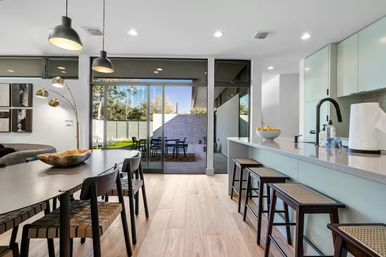 Sunlit modern open-plan kitchen and dining area with breakfast island and bar stools, pendant lights over a woven-seat dining table, light hardwood floors, and large sliding glass doors leading to a backyard patio with outdoor dining set.