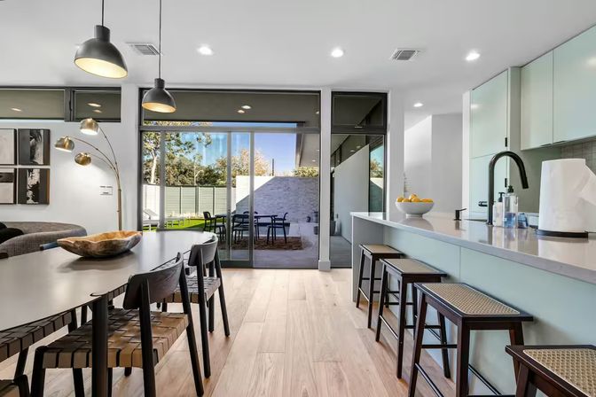 Sunlit modern open-plan kitchen and dining area with breakfast island and bar stools, pendant lights over a woven-seat dining table, light hardwood floors, and large sliding glass doors leading to a backyard patio with outdoor dining set.