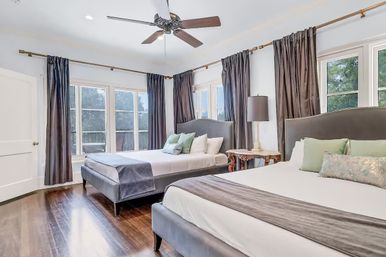 Spacious sunlit bedroom with two upholstered beds, large corner windows with dark drapes, ceiling fan and hardwood floors