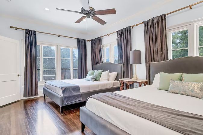 Spacious sunlit bedroom with two upholstered beds, large corner windows with dark drapes, ceiling fan and hardwood floors