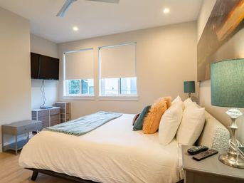 Bright modern apartment bedroom interior with queen bed, white linens, colorful accent pillows and teal lamp, wall-mounted TV, two windows with roller shades and light wood flooring.