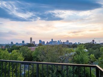 Balcony railing framing the Austin skyline at sunset, leafy treetops below and soft pastel clouds above.