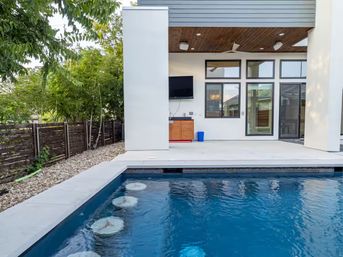 Modern backyard pool and covered patio with submerged bar stools, wall-mounted TV, ceiling fan, compact outdoor kitchenette, sliding glass doors, gravel landscaping and wood privacy fence with bamboo trees