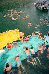 Summer lake party with a cheerful group floating on large colorful inflatable mats and swimming near a pontoon boat, socializing and holding drinks on green water.
