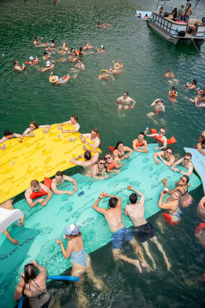 Summer lake party with a cheerful group floating on large colorful inflatable mats and swimming near a pontoon boat, socializing and holding drinks on green water.