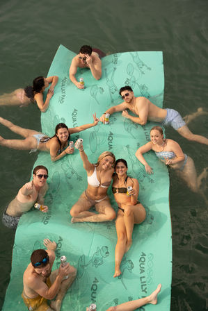 Group of friends lounging and cheering on a large aqua floating mat in a lake, holding canned drinks and smiling on a sunny summer day.