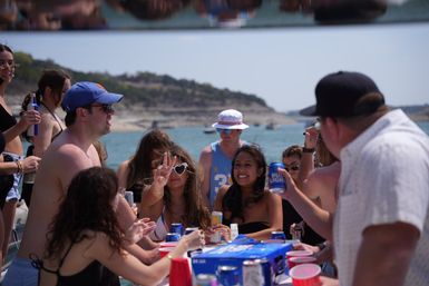 Sunlit summer boat party on a lake — friends laughing and raising beer cans and red cups aboard a pontoon with a rocky shoreline in the background.