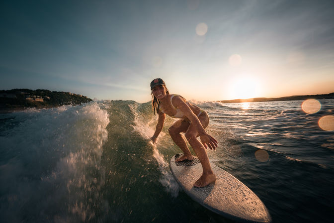 Surfer in a cap riding a breaking wave on a surfboard at sunset near a rocky coastline with golden sun flare over the ocean