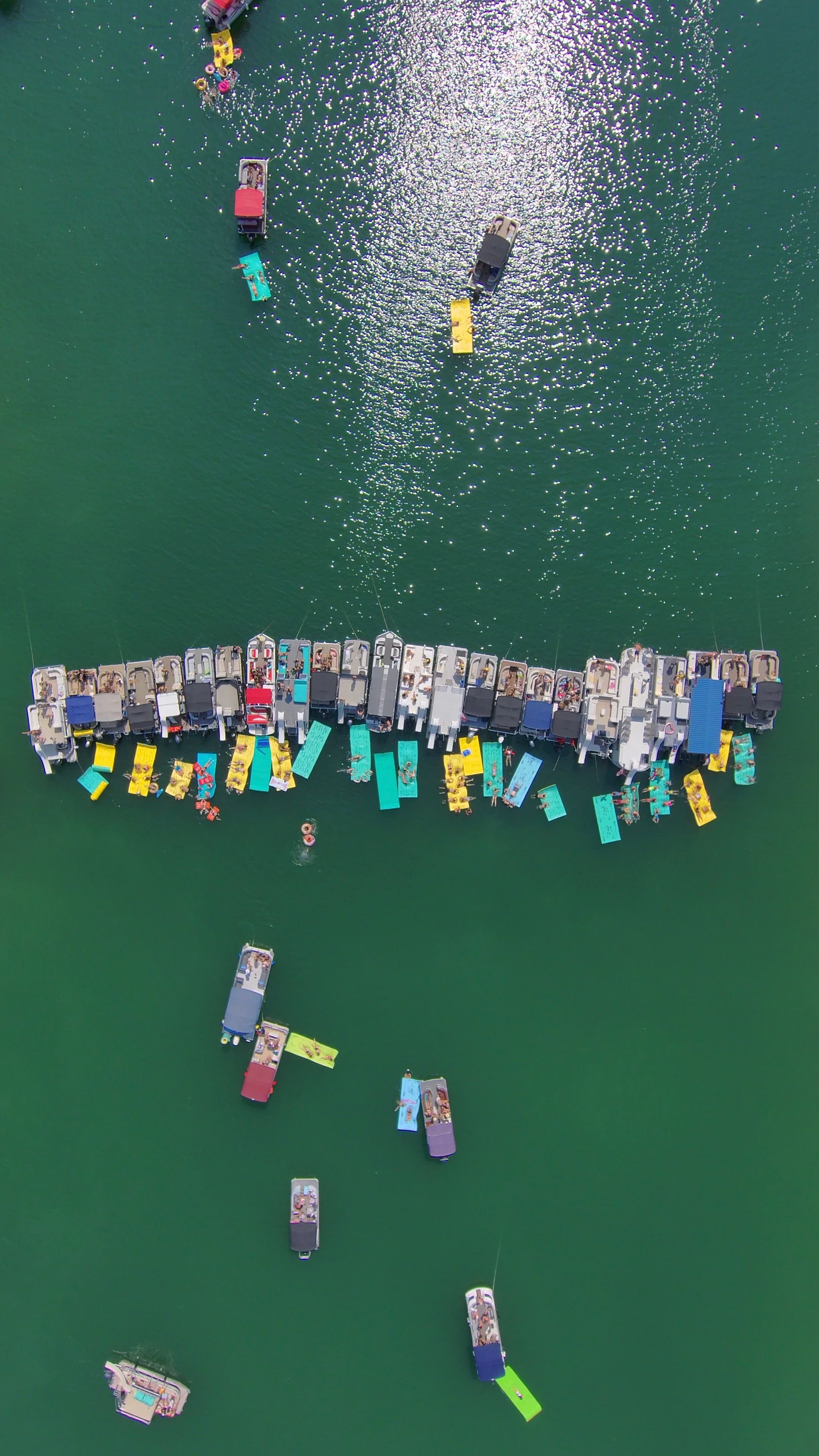 Aerial shot of a busy summer lake: a long dock lined with pontoon boats and bright yellow and teal floating pads, sun sparkling on emerald water with swimmers and anchored boats