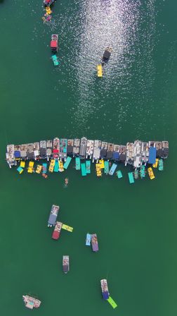 Aerial shot of a busy summer lake: a long dock lined with pontoon boats and bright yellow and teal floating pads, sun sparkling on emerald water with swimmers and anchored boats
