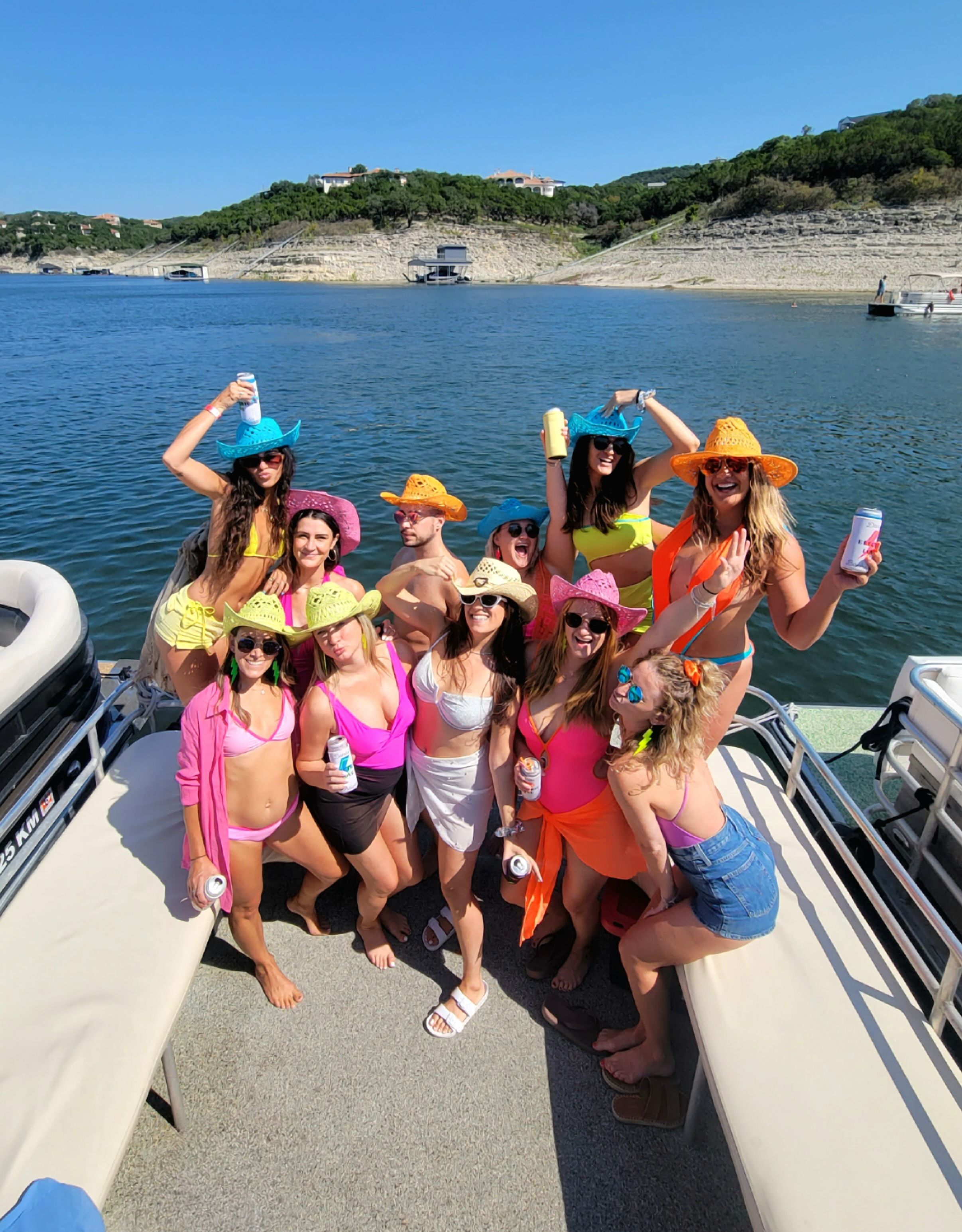 Group of friends on a pontoon boat lake party in colorful swimsuits and neon cowboy hats, holding drinks with rocky shoreline and lakeside homes in the background